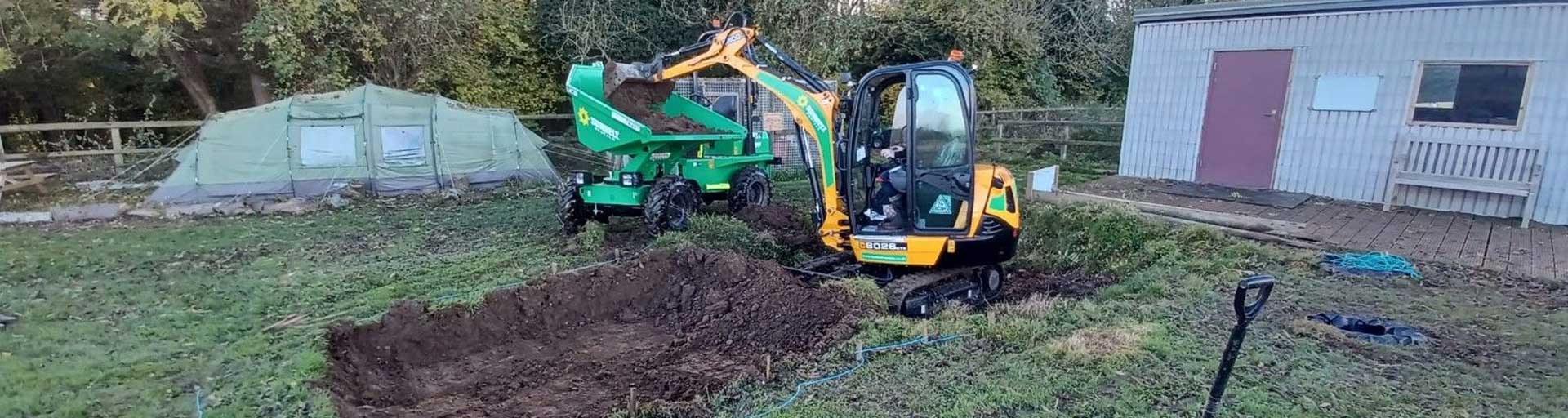 Mini Digger Unloading Dirt Into A Dumper On Patch Of Grass Worksite